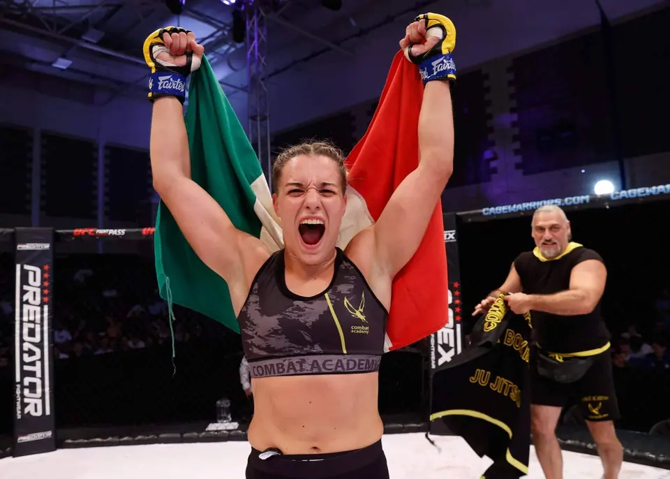 Un’atleta Combat Academy urla di gioia con il tricolore alzato dopo una vittoria in gabbia; il Maestro è alle sue spalle